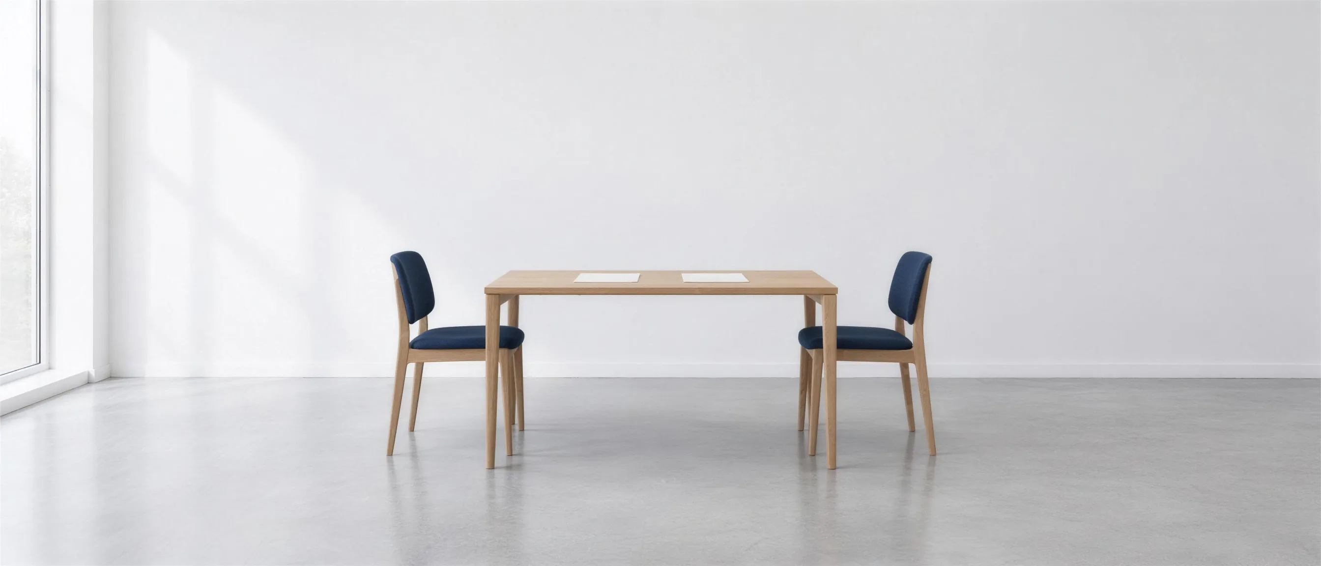 A light oak table with two sheets of paper on it and a chair with dark blue upholstery on either side, in a bright room