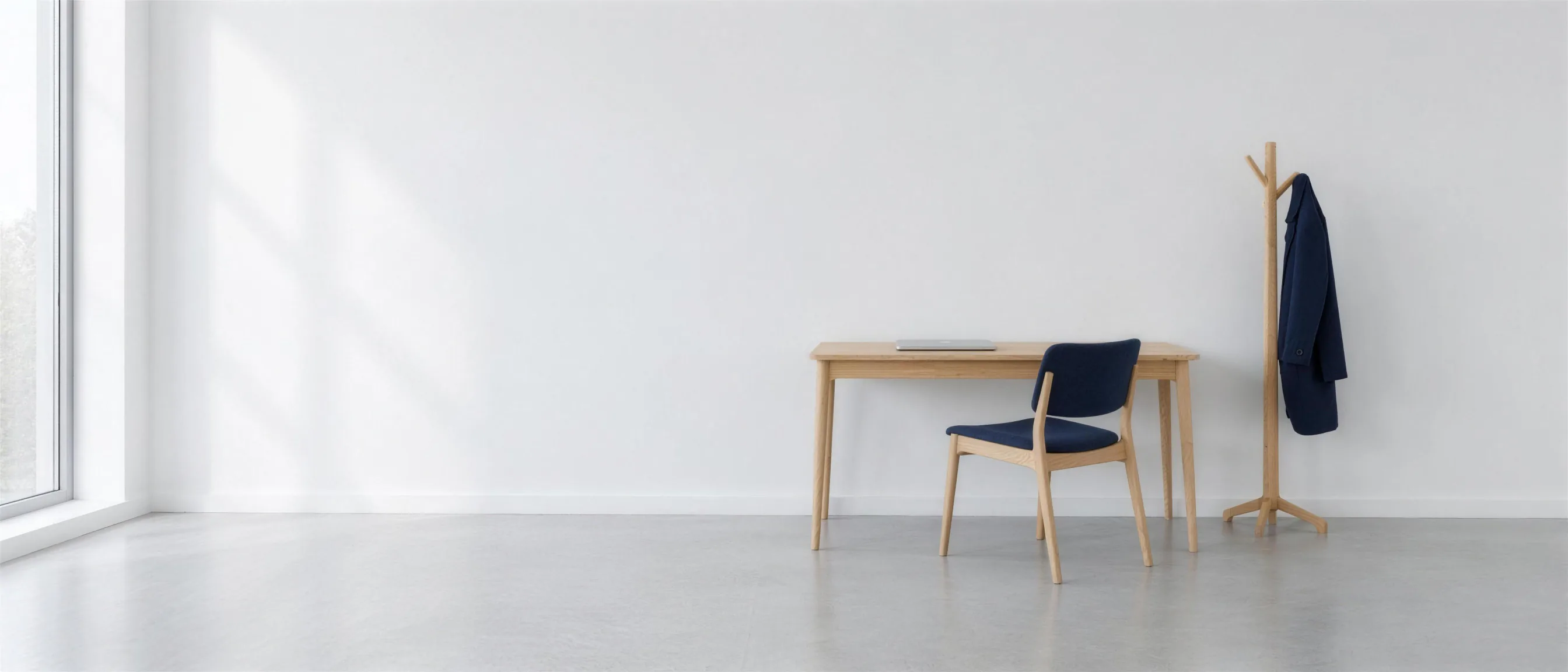 A light oak desk with a chair in a bright room, beside it a coat stand with a dark blue coat