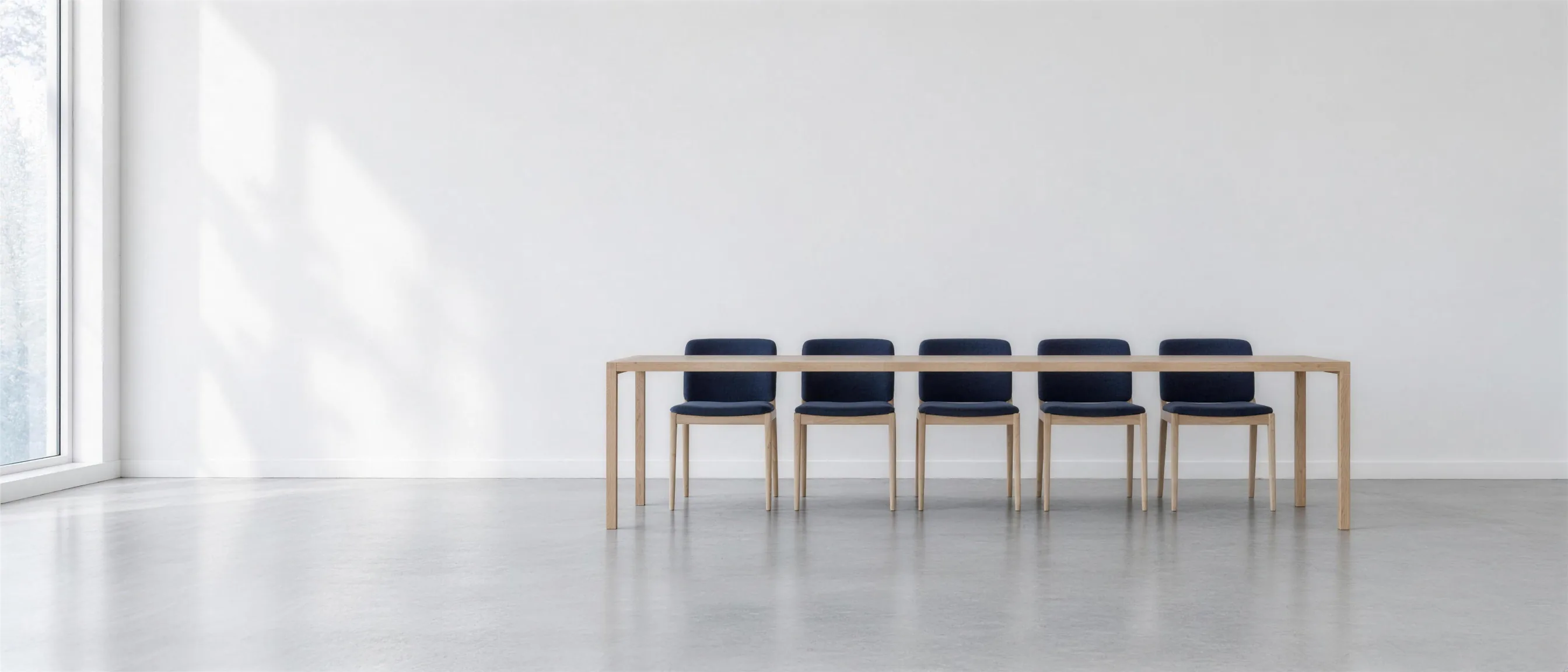 Five chairs with dark blue upholstery in a row at a long light oak table, in a bright room